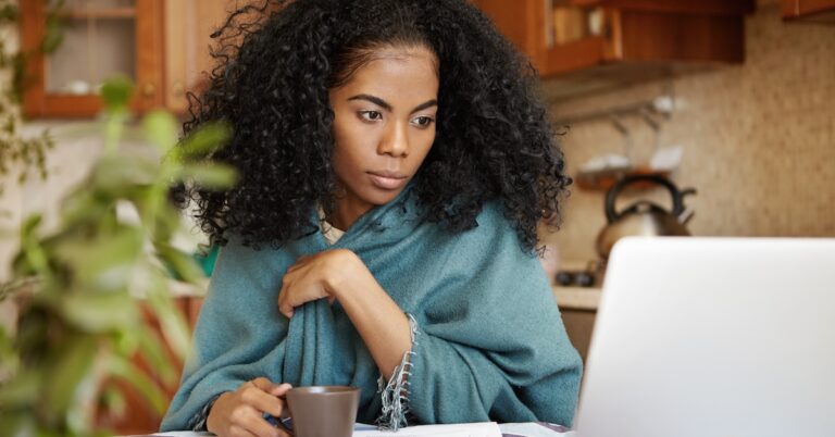 african american woman sitting at table drinking coffee while looking at laptop screen